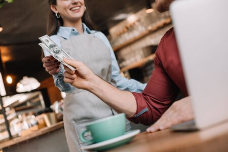 cropped view of freelancer with laptop giving cash to smiling waitress in coffee shopの写真素材