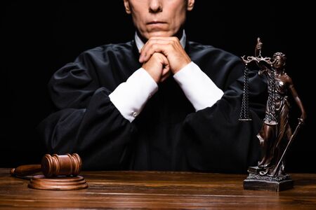 cropped view of judge in judicial robe sitting at table with gavel and themis figure isolated on blackの写真素材