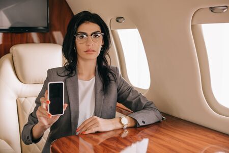 attractive businesswoman in glasses holding smartphone with blank screen in private jetの写真素材