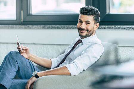 selective focus of handsome businessman smiling at camera while sitting in armchair and holding smartphoneの写真素材