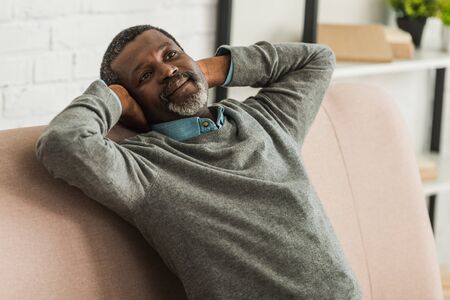 dreamy, positive african american man looking away while sitting on sofaの写真素材