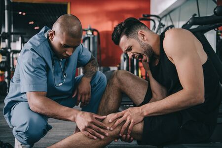 african american doctor examining injured knee of screaming sportsman