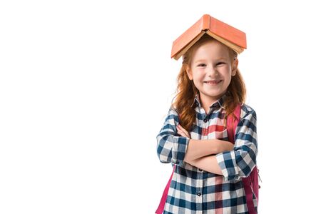 cheerful redhead pupil with orange book on head standing with crossed arms isolated on whiteの写真素材