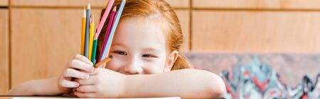panoramic shot of smiling redhead child holding color pencils