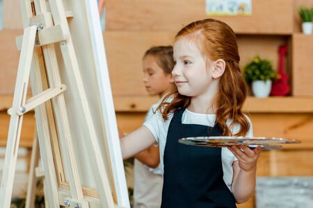 selective focus of cheerful redhead kid holding palette near easel and child in art schoolの写真素材