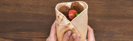 cropped view of woman holding falafel with vegetables and sauce in pita on wooden table, panoramic...
