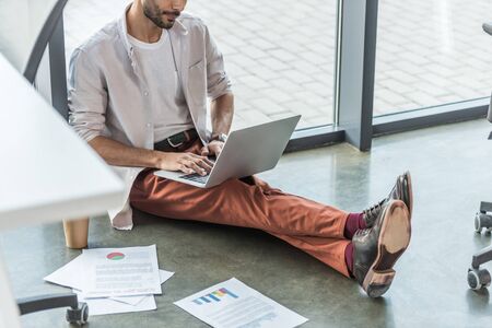 cropped view of businessman in casual clothes sitting on floor near documents and using laptopの写真素材