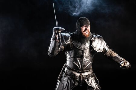 handsome and angry knight in armor fighting with sword on black backgroundの写真素材