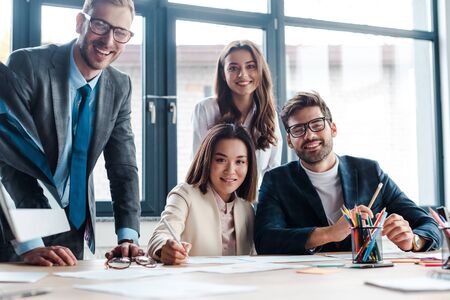 selective focus of happy businessmen and multicultural businesswoman looking at camera in office