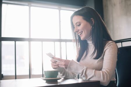 happy young woman sitting at wooden table with cappuccino and using smartphone on balcony in coffee...