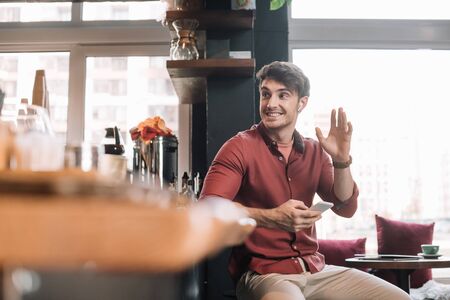 smiling handsome man sitting near bar counter in wireless earphones with smartphone and waving handの写真素材