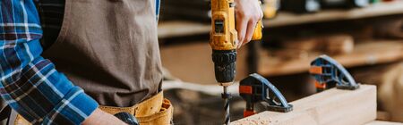 panoramic shot of carpenter in apron holding hammer drill near wooden plank