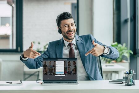 KYIV, UKRAINE - OCTOBER 10, 2019: Cheerful call center operator winking at camera and pointing with fingers at laptop with Linkedin website on screen.のeditorial素材