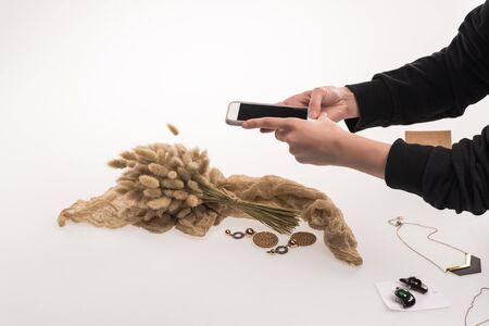 cropped view of photographer taking pictures of composition with flora and jewelry on smartphone on...