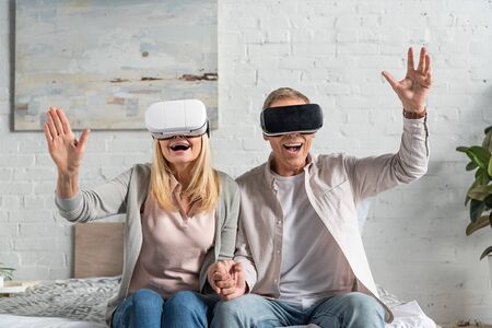 Exited couple in virtual reality headsets holding hands on bedの写真素材