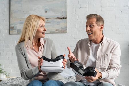 Exited couple holding virtual reality headsets on bed