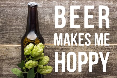 top view of fresh beer in bottle with green hop on wooden surface with beer makes me hoppy illustrationの写真素材
