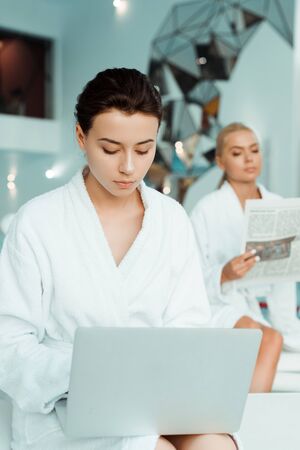 selective focus of attractive woman using laptop and friend reading newspaper on background in spaの写真素材