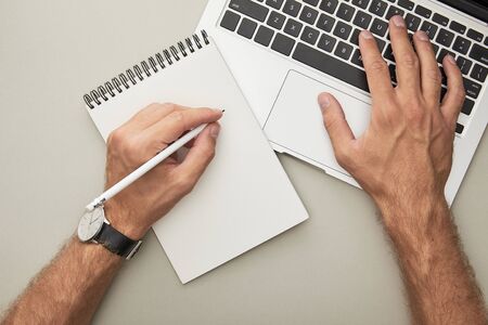 cropped view of man using laptop while holding pencil near notebook isolated on grey