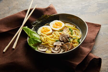 traditional spicy ramen in bowl with chopsticks on brown napkin on stone surface