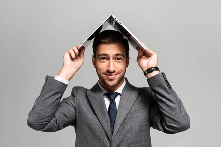 handsome and smiling businessman in suit holding laptop above head isolated on grey