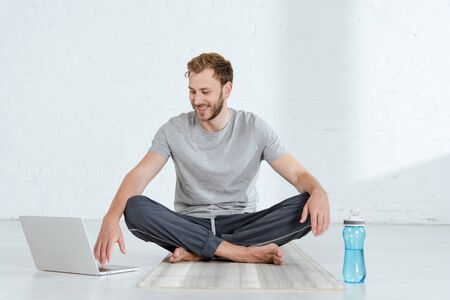 smiling man sitting in easy pose near laptop and sports bottle