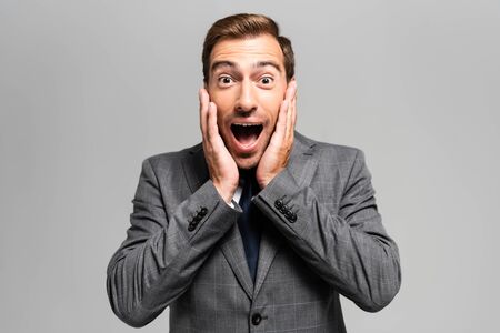 handsome and shocked businessman in suit looking at camera isolated on grey