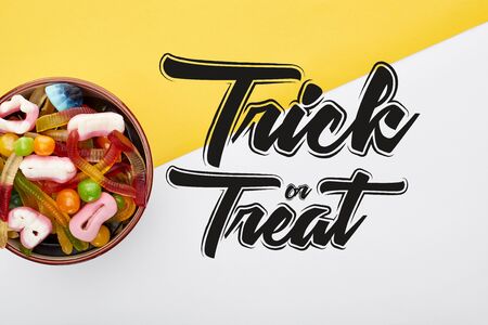 top view of colorful gummy sweets in bowl on yellow and white background with trick or treat illustrationの写真素材