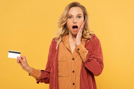 Shocked blonde woman holding credit card isolated on yellow
