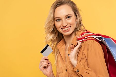 Smiling woman holding credit card and shopping bags isolated on yellow