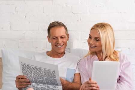 Man reading newspaper to wife with digital tablet on bedの写真素材