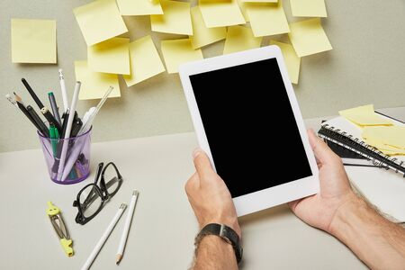 cropped view of man holding digital tablet with blank screen near stationery and notebooks on grey...