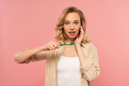 Shocked blonde woman holding toothbrush isolated on pink