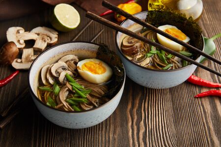 traditional spicy ramen in bowls with chopsticks and vegetables on wooden table with napkinの写真素材