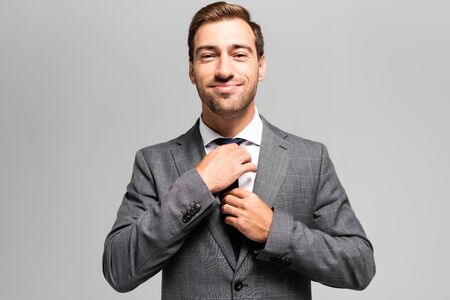 handsome and smiling businessman in suit touching tie isolated on grey