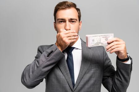 handsome businessman in suit obscuring face and holding dollar banknotes isolated on grey