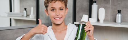 panoramic shot of happy boy holding bottle with green mouthwash and showing thumb up in bathroom
