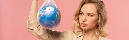 Panoramic shot of blonde woman looking at globe in polyethylene pack isolated on pink, global...