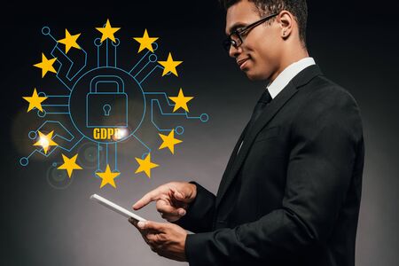 side view of smiling african american businessman using digital tablet on dark background with gdpr illustrationの写真素材