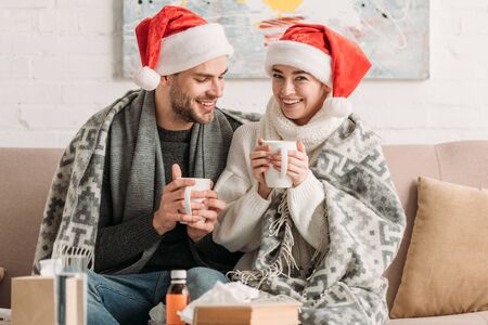 diseased, smiling couple in santa hats, covered with blanket, holding cups of warming drink while sitting on sofaの写真素材