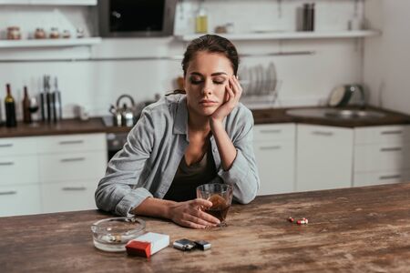 Worried woman holding whiskey glass beside cigarettes and pills on tableの写真素材