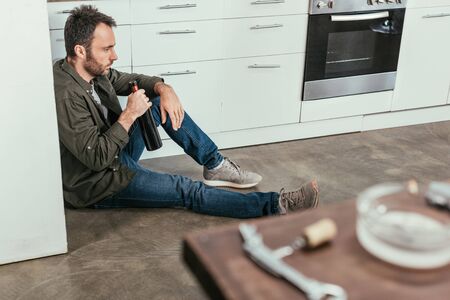 Selective focus of sad man with wine bottle sitting on kitchen floorの写真素材