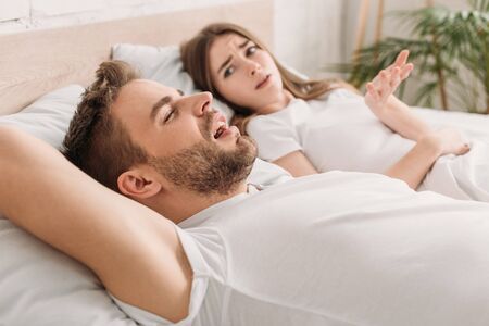dissatisfied woman showing outrage gesture while lying in bed near snoring husband
