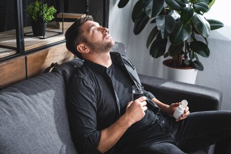 handsome man with panic attack holding pills and glass of water in apartmentの写真素材