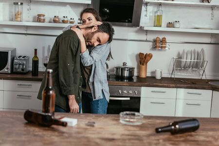 Selective focus of woman calming down worried husband with alcohol addiction at kitchenの写真素材
