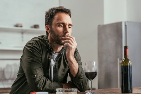 Upset man smoking cigarette beside wine on tableの写真素材