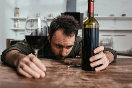 Selective focus of sad man holding wine glass and bottle on kitchen tableの写真素材