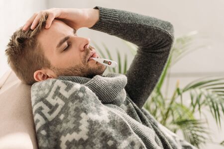 sick man, wrapped in blanket, sitting on sofa with closed eyes and measuring temperatureの写真素材