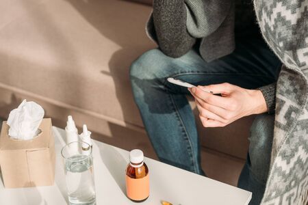 cropped view of sick man sitting near table with medicines and holding thermometerの写真素材