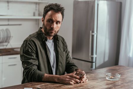 Worried man holding whiskey glass beside ashtray on kitchen table
